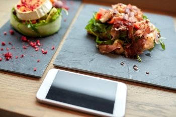 food, eating and technology concept - goat cheese and prosciutto ham salads on stone plates with smartphone at restaurant or cafe