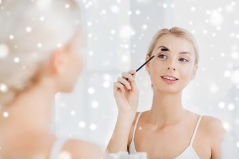beauty, make up, morning and people concept - young woman brushing eyebrow with brush and looking to mirror at home bathroom over snow