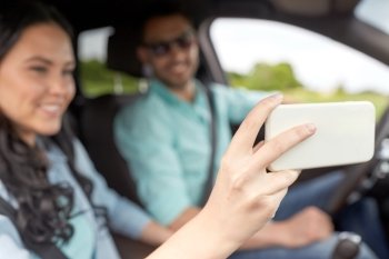 technology, travel, vacation, road trip and people concept - happy man and woman driving in car and taking selfie with smartphone
