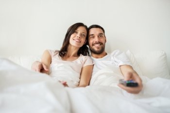 people, rest, love, relationships and television concept - happy couple with remote lying in bed at home and watching tv