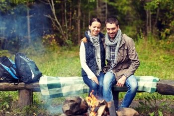 camping, travel, tourism, hike and people concept - happy couple sitting on bench and warming near campfire at camp in woods