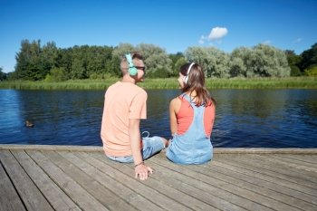 holidays, vacation, love and people concept - happy teenage couple with headphones sitting on river berth and listening to music at summer