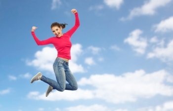 happiness, freedom, power, motion and people concept - smiling young woman jumping in air with raised fists over blue sky and clouds background