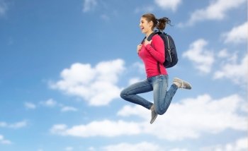 education, travel, tourism, motion and people concept - smiling young woman or student with backpack jumping in air over blue sky and clouds background