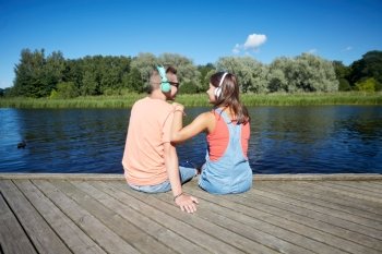 holidays, vacation, love and people concept - happy teenage couple with headphones sitting on river berth and listening to music at summer