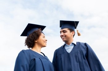 education, graduation and people concept - happy international students in mortarboards and bachelor gowns outdoors. happy students or bachelors in mortarboards