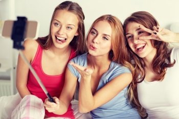 friendship, people, pajama party and technology concept - happy friends or teenage girls with smartphone and monopod taking selfie at home. teen girls with smartphone taking selfie at home