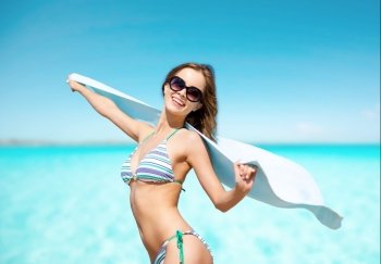 people, summer holidays and vacation concept - beautiful woman in bikini and sunglasses with towel on beach over blue sky and sea background. woman in bikini and sunglasses with towel on beach
