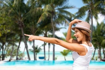 summer holidays, travel, people and vacation concept - happy young woman in hat pointing finger over exotic tropical beach with palm trees and pool background. happy young woman in hat on summer beach
