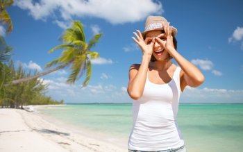 summer holidays, travel, people and vacation concept - happy young woman in hat making finger glasses over exotic tropical beach with palm trees and sea shore background. happy young woman in hat on summer beach