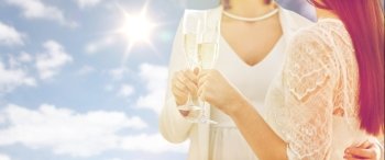 people, homosexuality, same-sex marriage, celebration and love concept - close up of happy married lesbian couple holding and clinking champagne glasses over sky and sun background. close up of lesbian couple with champagne glasses