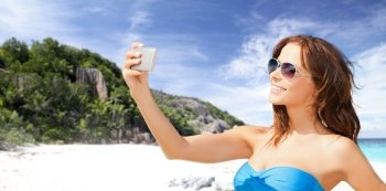 technology, summer holidays, travel and people concept - happy young woman in bikini swimsuit and sunglasses taking selfie with smatphone over beach and palm trees background. woman in swimsuit taking selfie with smatphone