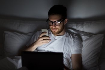technology, internet, communication and people concept - young man in glasses with laptop computer and smartphone in bed at home bedroom at night. man with laptop and smartphone at night in bed