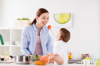 family, food, healthy eating, cooking and people concept - little baby feeding his mother with carrot at home kitchen. baby feeding mother with carrot at home kitchen