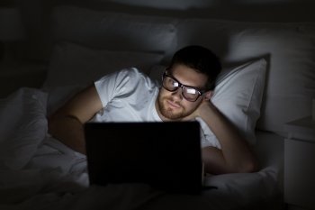 technology, internet, communication and people concept - young man in glasses with laptop computer in bed at home bedroom at night. young man with laptop in bed at home bedroom