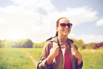 travel, hiking, backpacking, tourism and people concept - happy young woman in sunglasses with backpack walking along country road outdoors. happy young woman with backpack hiking outdoors
