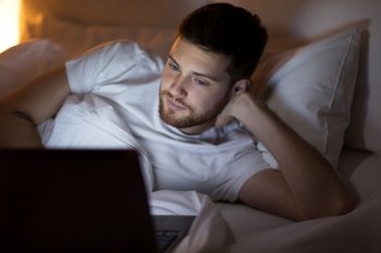 technology, internet, communication and people concept - young man with laptop computer in bed at home bedroom at night. young man with laptop in bed at home bedroom
