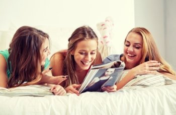 friendship, people and pajama party concept - happy friends or teenage girls reading magazine in bed at home. friends or teen girls reading magazine at home