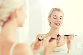 beauty, make up, cosmetics, morning and people concept - young woman applying eyeshade with makeup brush and looking to mirror at home bathroom. woman with makeup brush and eyeshade at bathroom