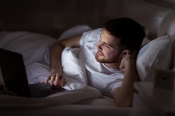 technology, internet, communication and people concept - happy smiling young man with laptop computer in bed at home bedroom at night. happy young man with laptop in bed at home