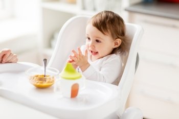 food, child, feeding and people concept - happy little baby girl with puree sitting in highchair and eating at home. happy baby girl with food and drink eating at home