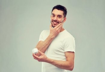 beauty, skin care, body care and people concept - smiling young man applying cream to face over gray background. happy young man applying cream to face over gray