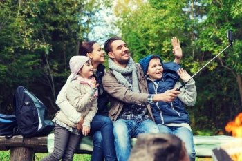 camping, travel, tourism, hike and people concept - happy family sitting on bench and taking picture with smartphone on selfie stick at campfire in woods. family with smartphone taking selfie near campfire. family with smartphone taking selfie near campfire