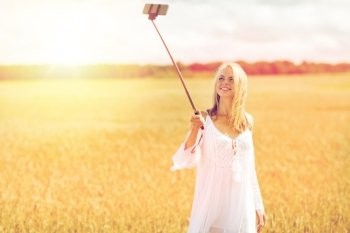 technology, summer holidays, vacation and people concept - smiling young woman in white dress taking picture by smartphone selfie stick on cereal field. happy young woman taking selfie by smartphone. happy young woman taking selfie by smartphone