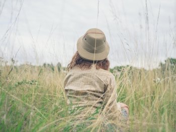 A young woman wearing a safari hat is sitting in the tall grass in a wilderness