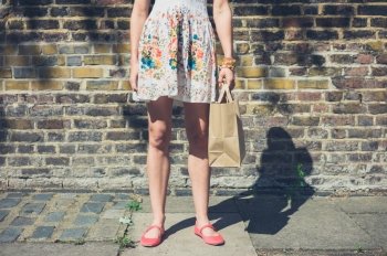 A young woman wearing a summer dress is standing in the street with a paper bag