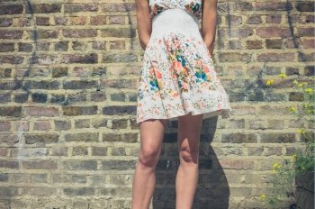 A young woman wearing a summer dress is standing by a brick wall outside on a sunny day
