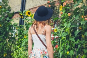 A young woman wearing a hat and a dress is admiring some sunflowers outside a house on a sunny day in summer