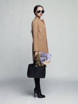 Fashion studio photo of young stylish woman. Beige coat, black leather boots and bag, bouquet of lavender. Catalogue clothes. Lookbook