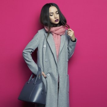 Fashion studio photo of young stylish woman on fuchsia background. Gray coat, pink scarf, purple lipstick, leather bag, . Catalogue clothes and accessories. Lookbook