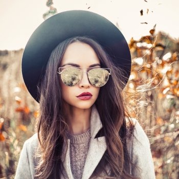 Outdoor fashion photo of young beautiful lady in autumn landscape with dry flowers. Gray coat, black hat, sunglusses, wine lipstick. Warm Autumn. Warm Spring