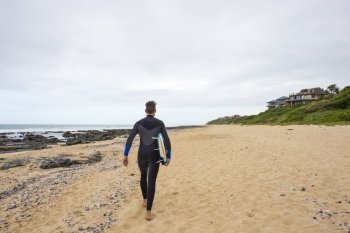A surfer in a wetsuit walks along the beach with his surfboard under his arm.