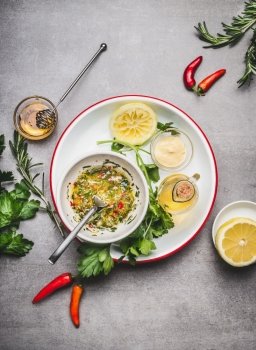 Homemade sauce or salad dressing in bowl with ingredients:  fresh herbs, oil, lemon and honey on gray concrete background, top view, flat lay. Healthy , clean food concept