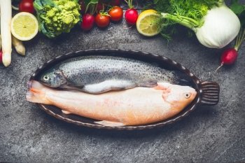 Cooking preparation of fish dishes with two raw trout and vegetables ingredients on grey concrete background, top view