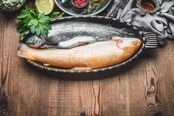 Two raw trout fishes in baking dish with ingredients on rustic wooden background, top view, cooking preparation