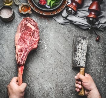 Male hands of  butcher or cook holding tomahawk beef steak and meat cleaver on dark rustic kitchen table background with cooking ingredients and condiment, top view, place for text