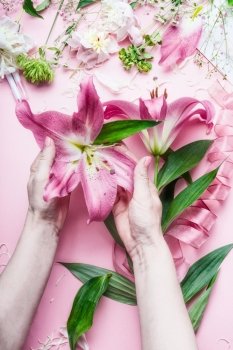 Creative Florist workspace. Female hands holding beautiful big pink lily flowers on pastell table with florist decoration equipment, top view, frame.Festive holiday concept