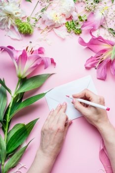 Female hands write with pencil greeting card on Blank envelop on pink table background with lily flowers and florist decoration equipment, top view. Creative  Invitation and holiday  concept