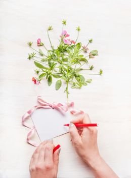 Female hand Write a greeting card on white wooden background with flowers bunch and pink ribbon, top view, copy space,  mock up