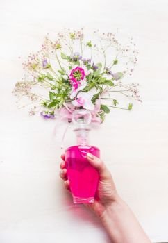 Female hand holding bottle with pink lotion and flowers and herbs bunch on white wooden background, top view. Organic herbal and  botanical cosmetic, Perfume and beauty concept