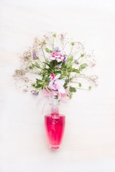 Cosmetic product glass bottle with pink liquid and bunch of flowers on white wooden background, top view.