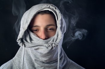 portrait of young man wearing a grey hood with half face covered and smoke around him against black background
