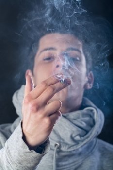 young man wearing a grey hood smoking a cigar with smoke around him against black background