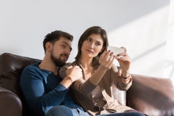 Cheerful couple taking a selfie with a smartphone at home sitting on couch