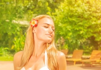 Closeup portrait of beautiful woman with closed eyes and red frangipani flower in hair, enjoying warm sunny day in fresh green garden, summer holiday concept