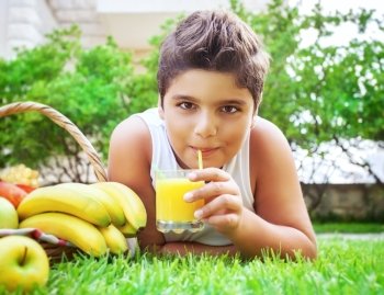 Portrait of a happy teen boy lying down on fresh green grass field and drinking tasty juice, eating fruits, enjoying healthy nutrition in the summer camp
